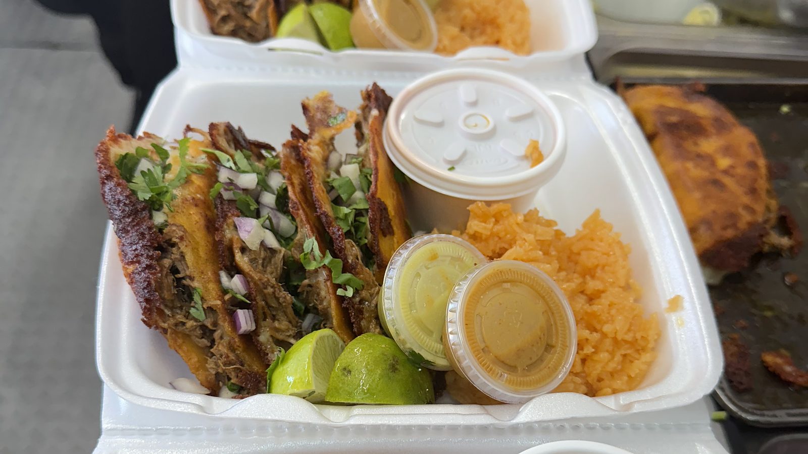 Photo from La TacoTrona's own Google listing: a row of open takeout trays on the counter, each with quesabirria tacos, limes, salsa cups and red rice.